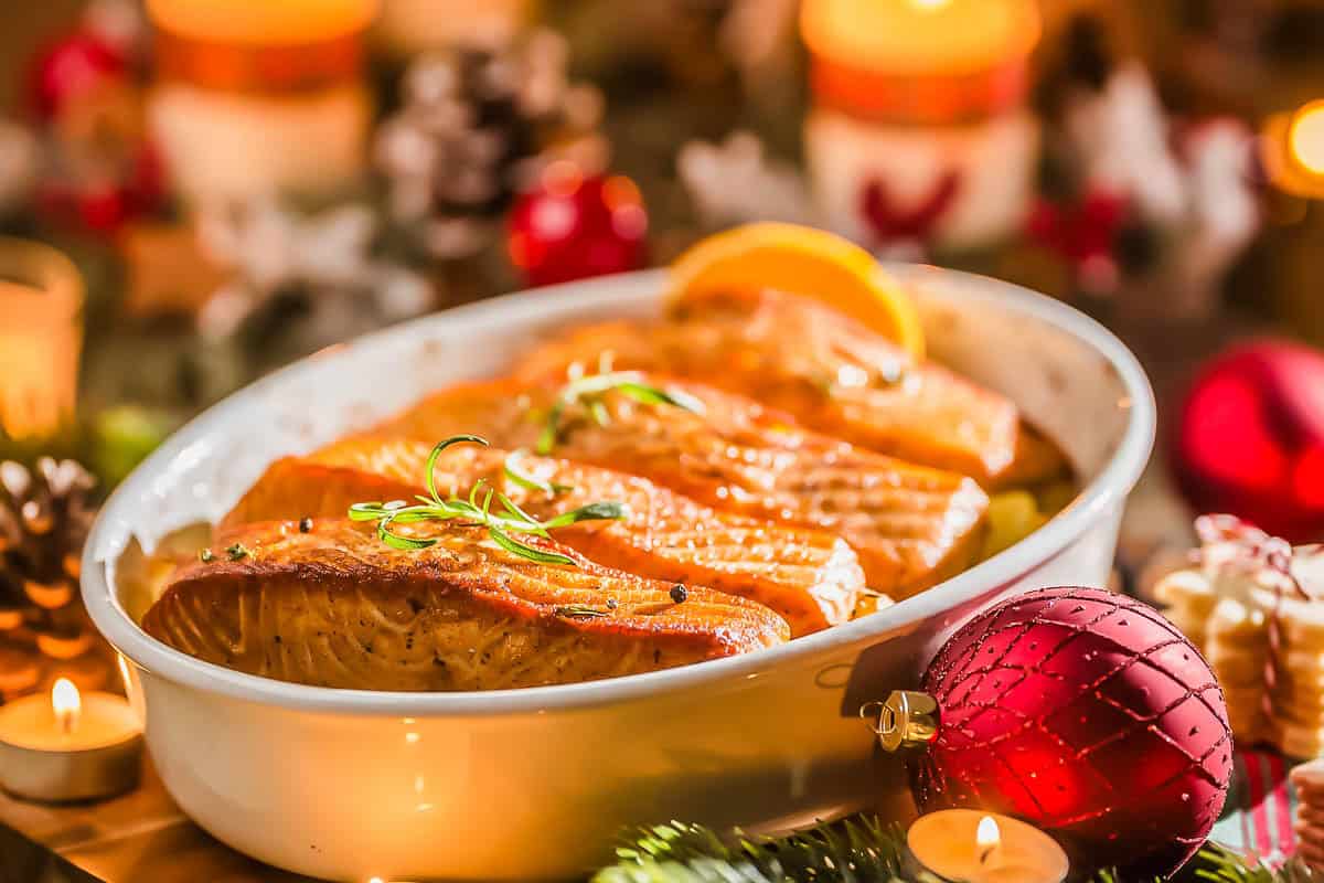 A festive dish of baked salmon fillets garnished with herbs is served in a white ceramic dish, surrounded by Christmas decorations, candles, pinecones, and ornaments.