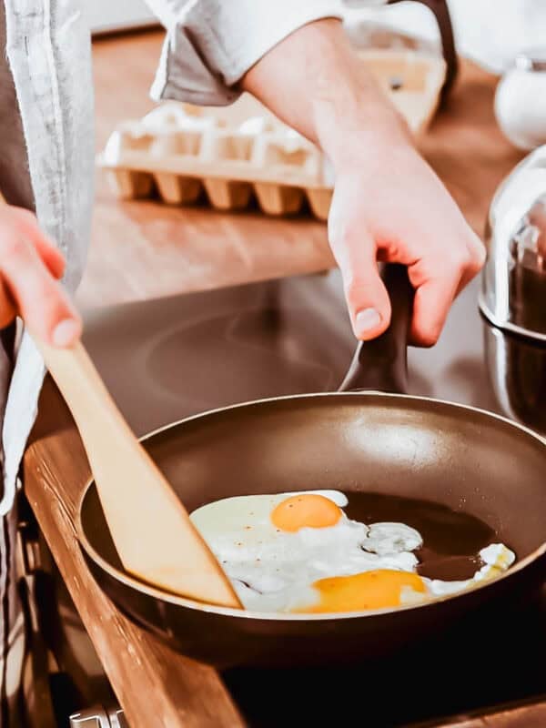 A person in pajamas cooks two eggs in a frying pan on a stovetop, stirring with a wooden spatula. A kettle and a carton of eggs are visible in the background.
