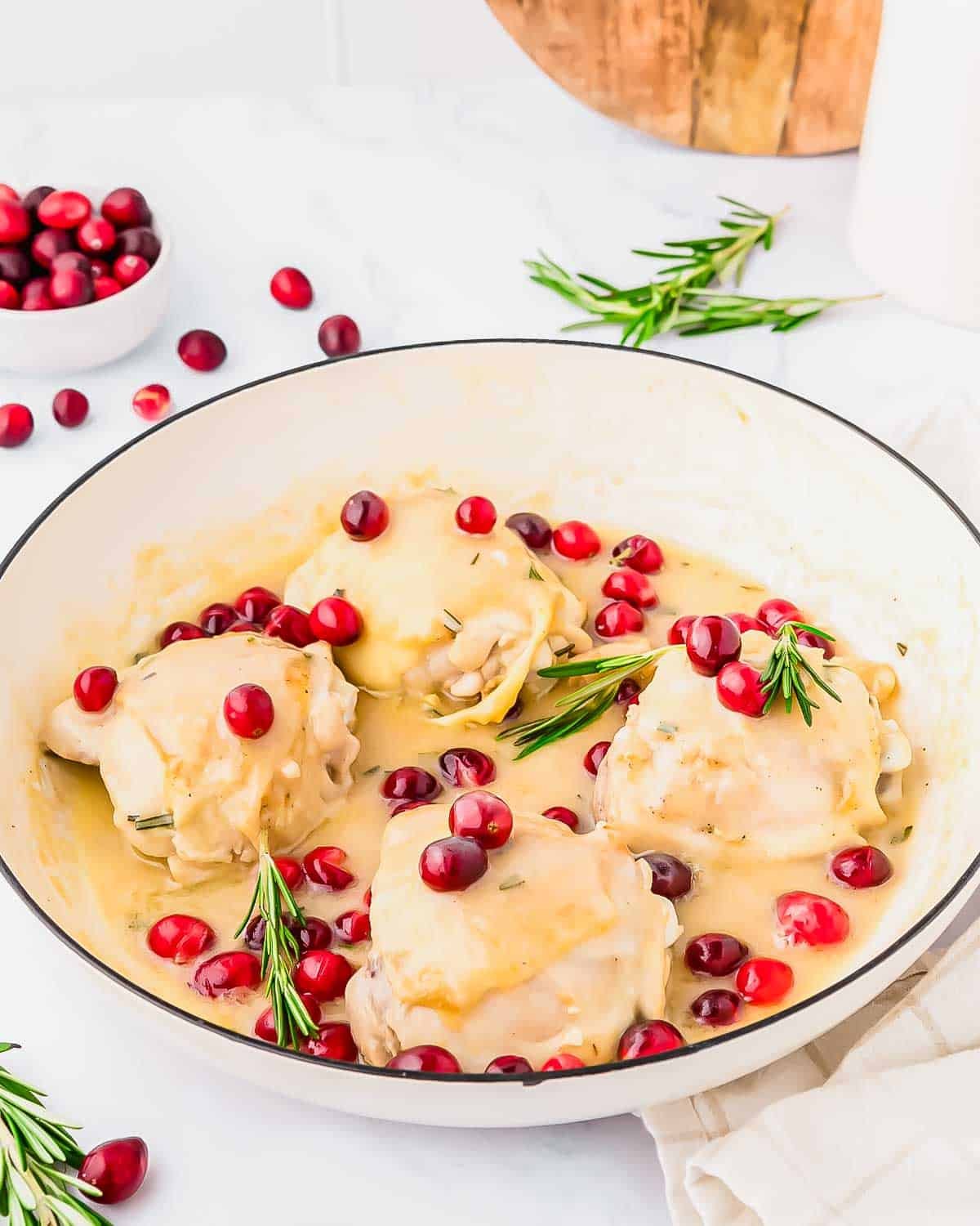 Chicken thighs in a creamy sauce white wine sauce are garnished with fresh rosemary sprigs and bright red cranberries in a white pan, with extra cranberries scattered around on a light countertop.