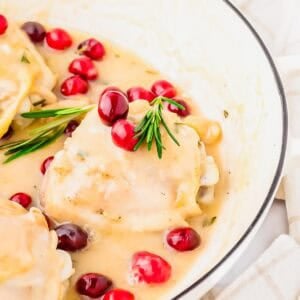 Chicken breasts in creamy white sauce, garnished with fresh cranberries and rosemary, served in a white dish with a beige-striped cloth nearby.