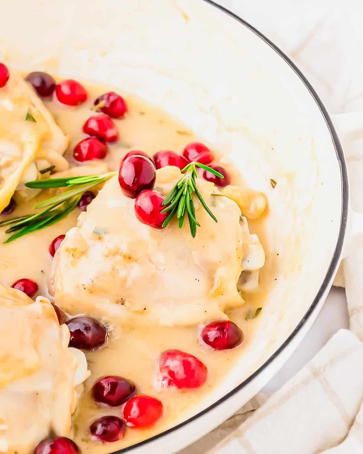 Chicken breasts in creamy white sauce, garnished with fresh cranberries and rosemary, served in a white dish with a beige-striped cloth nearby.