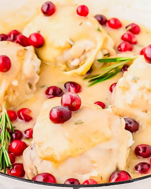 Four chicken thighs in creamy sauce are garnished with fresh cranberries and rosemary sprigs in a white dish. The sauce is thick and glossy, surrounding the chicken and vibrant cranberries.