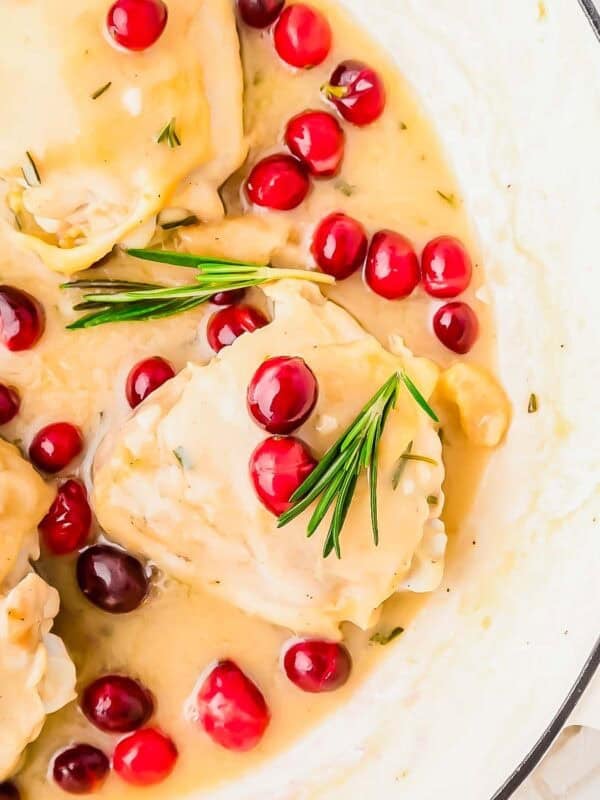 Chicken thighs with cranberries in a creamy sauce, garnished with fresh sprigs of rosemary, served in a white skillet.