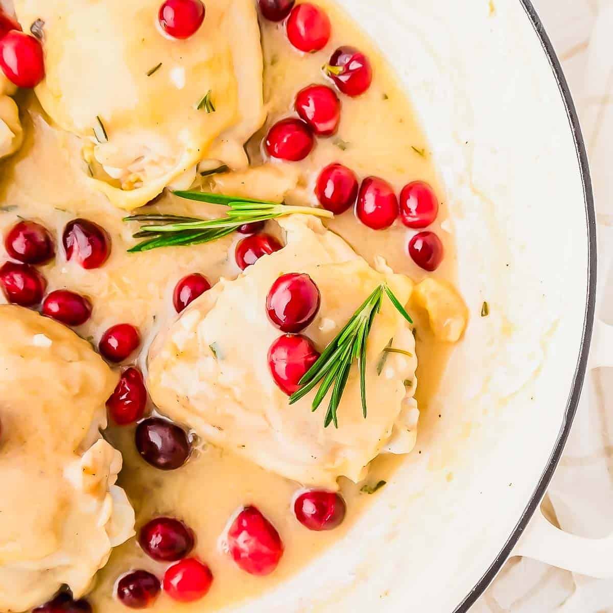 Chicken thighs with cranberries in a creamy sauce, garnished with fresh sprigs of rosemary, served in a white skillet.