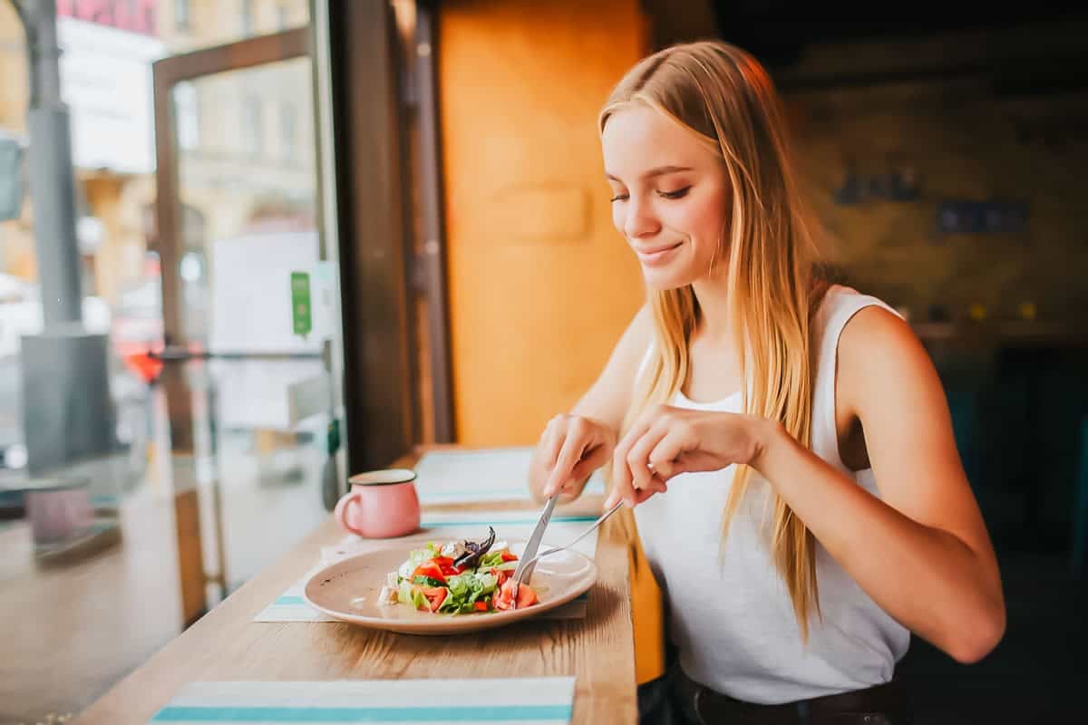 A young woman with long blonde hair sits at a wooden counter by a window, smiling as she cuts into a fresh salad on a plate. A pink mug sits beside her. Warm sunlight streams into the cozy cafe.