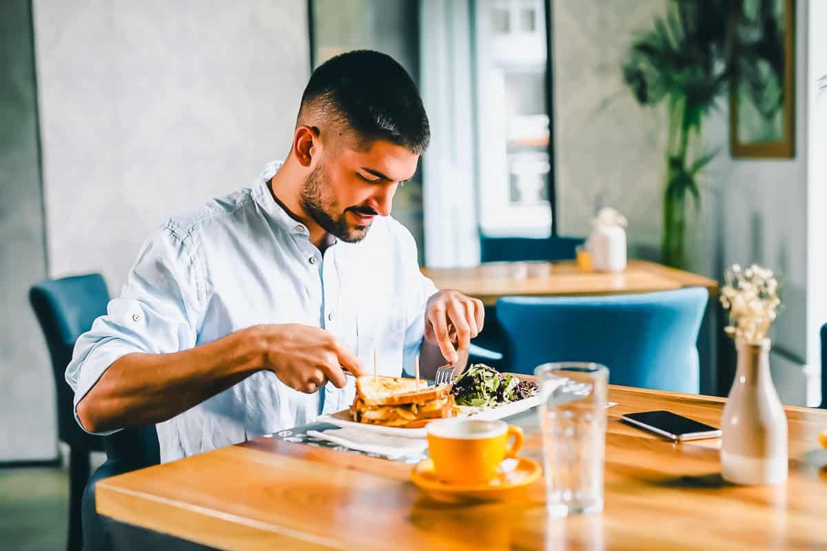 A man in a light blue shirt sits alone at a wooden table in a modern café, eating a meal with toast and salad. A yellow cup, glass of water, phone, and small flower vase are on the table.