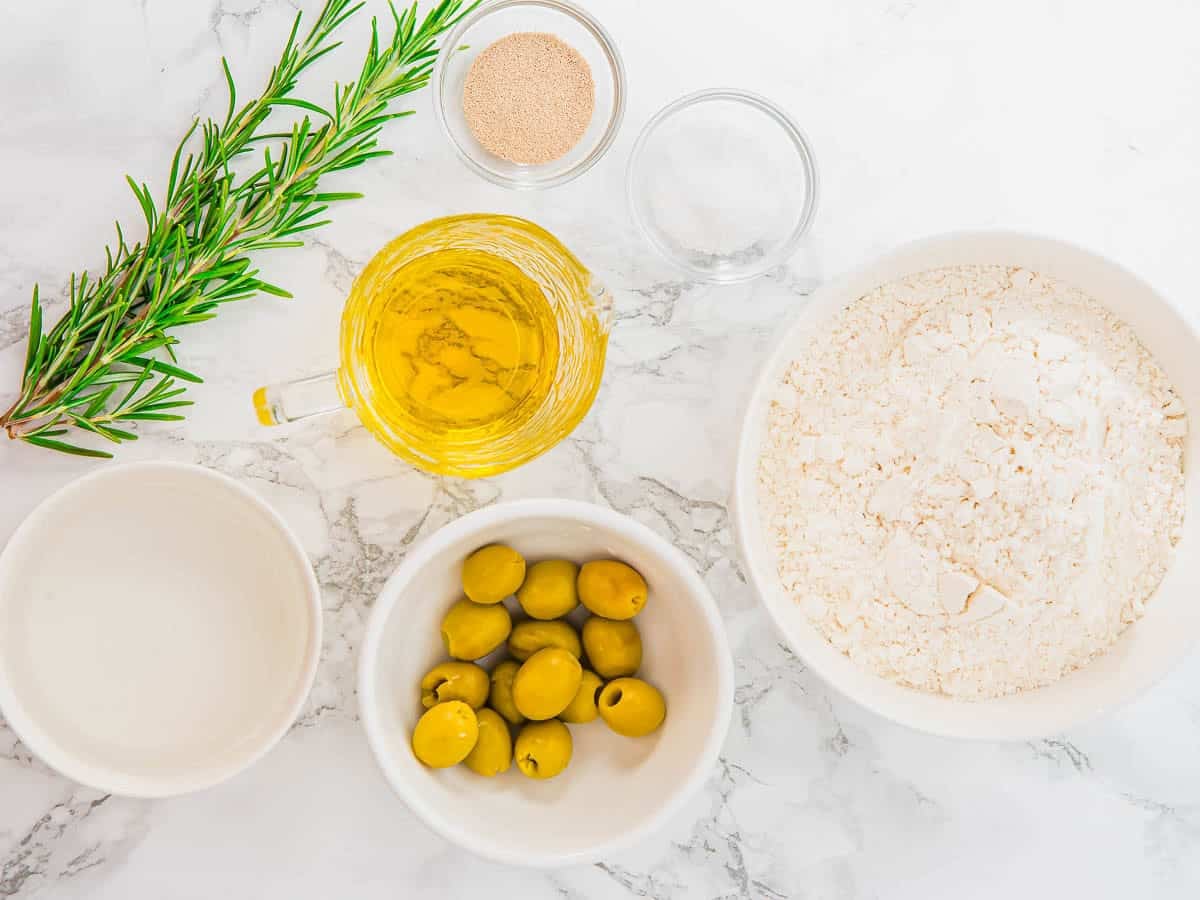 A top-down view of several ingredients on a marble surface: a bowl of flour, a bowl of green olives, a glass jug of olive oil, a small bowl of yeast, a bowl of water, and a sprig of fresh rosemary.