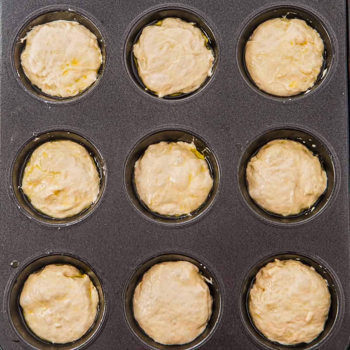 A muffin tin filled with nine muffin cups, each holding an even portion of focaccia dough, ready to rise. The batter appears light in color and slightly glossy.