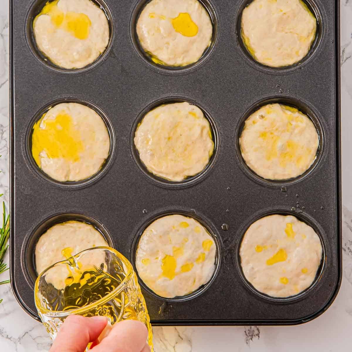 A hand pours olive oil from a glass onto unbaked focaccia muffin batter in a nonstick muffin pan, with each cup filled and drizzled with oil. The pan sits on a white marble countertop.
