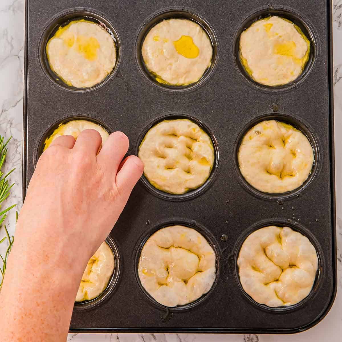 A hand presses dimples into dough rounds in a nonstick muffin tin, preparing mini focaccia breads. The dough is drizzled with olive oil. The muffin tin rests on a marble countertop.