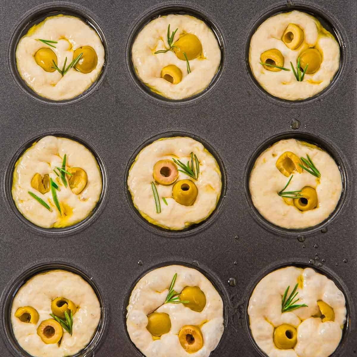A muffin tin filled with unbaked focaccia dough, each portion topped with sliced green olives and sprigs of fresh rosemary, ready to be baked.