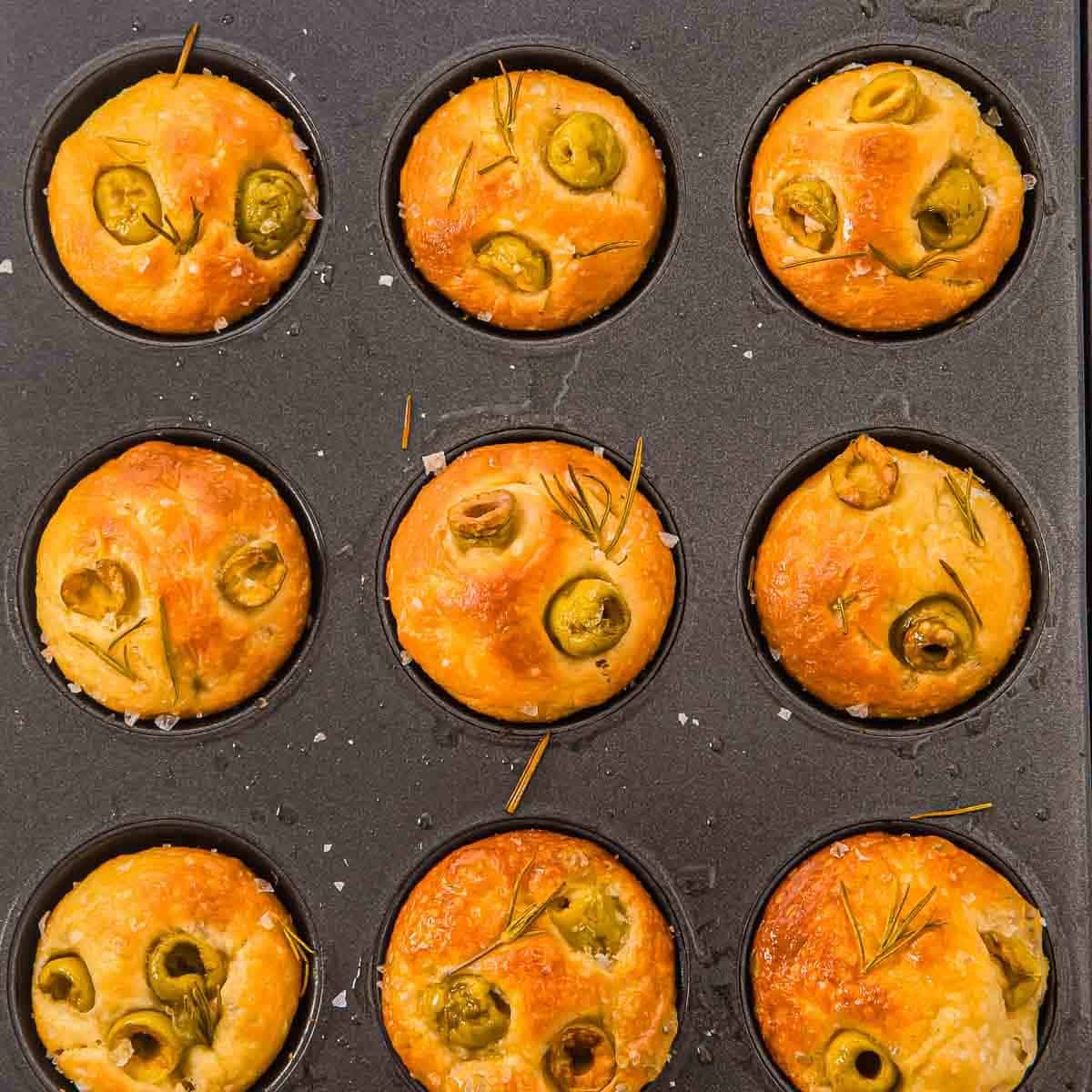 A muffin tin with nine baked focaccia muffin rolls topped with green olives, rosemary, and sea salt. The golden-brown rolls are evenly placed in each cup of the dark metal pan.