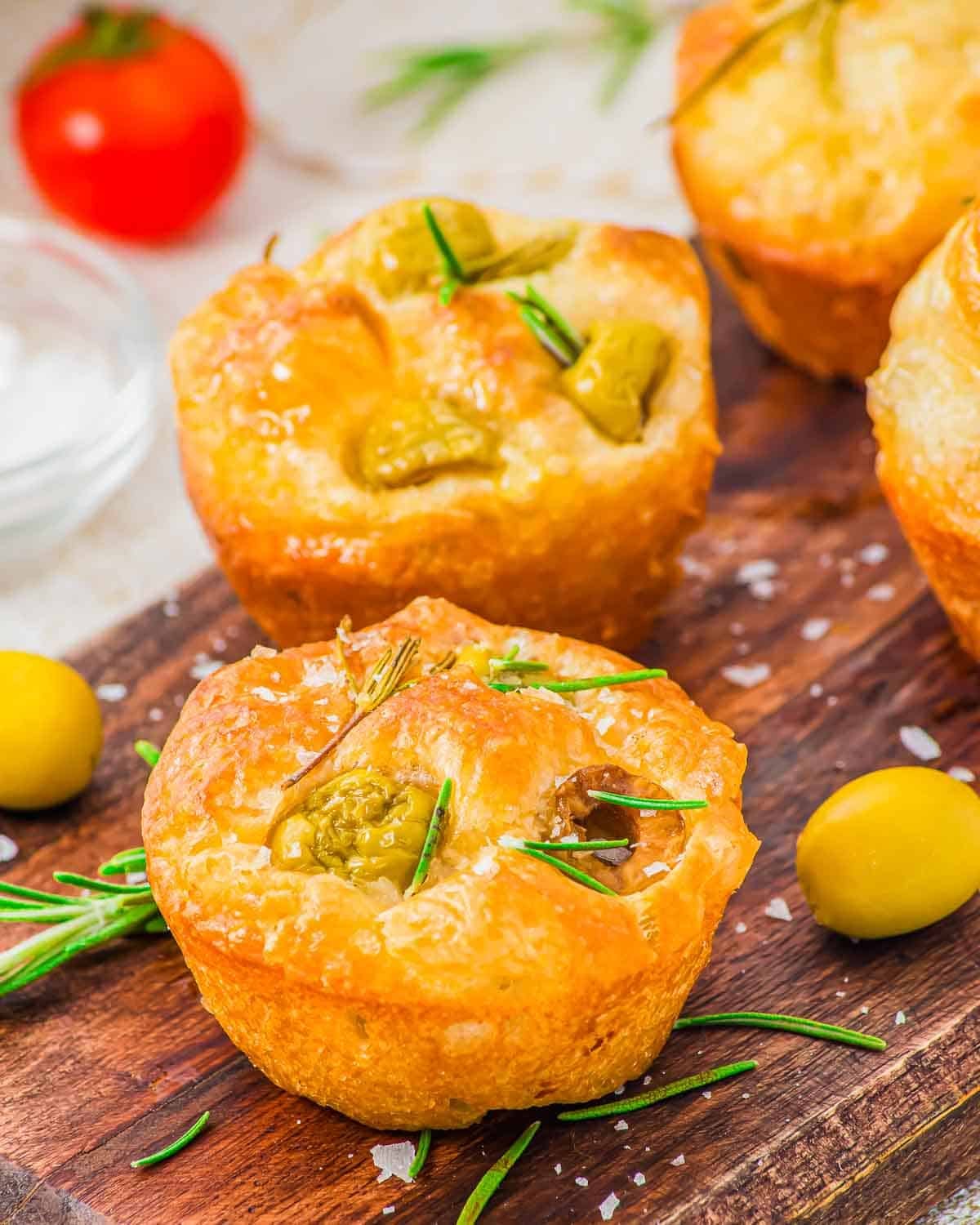 Golden baked focaccia muffins topped with green olives and rosemary sit on a wooden board, with scattered sea salt, rosemary sprigs, and yellow olives around them. A cherry tomato is visible in the background.