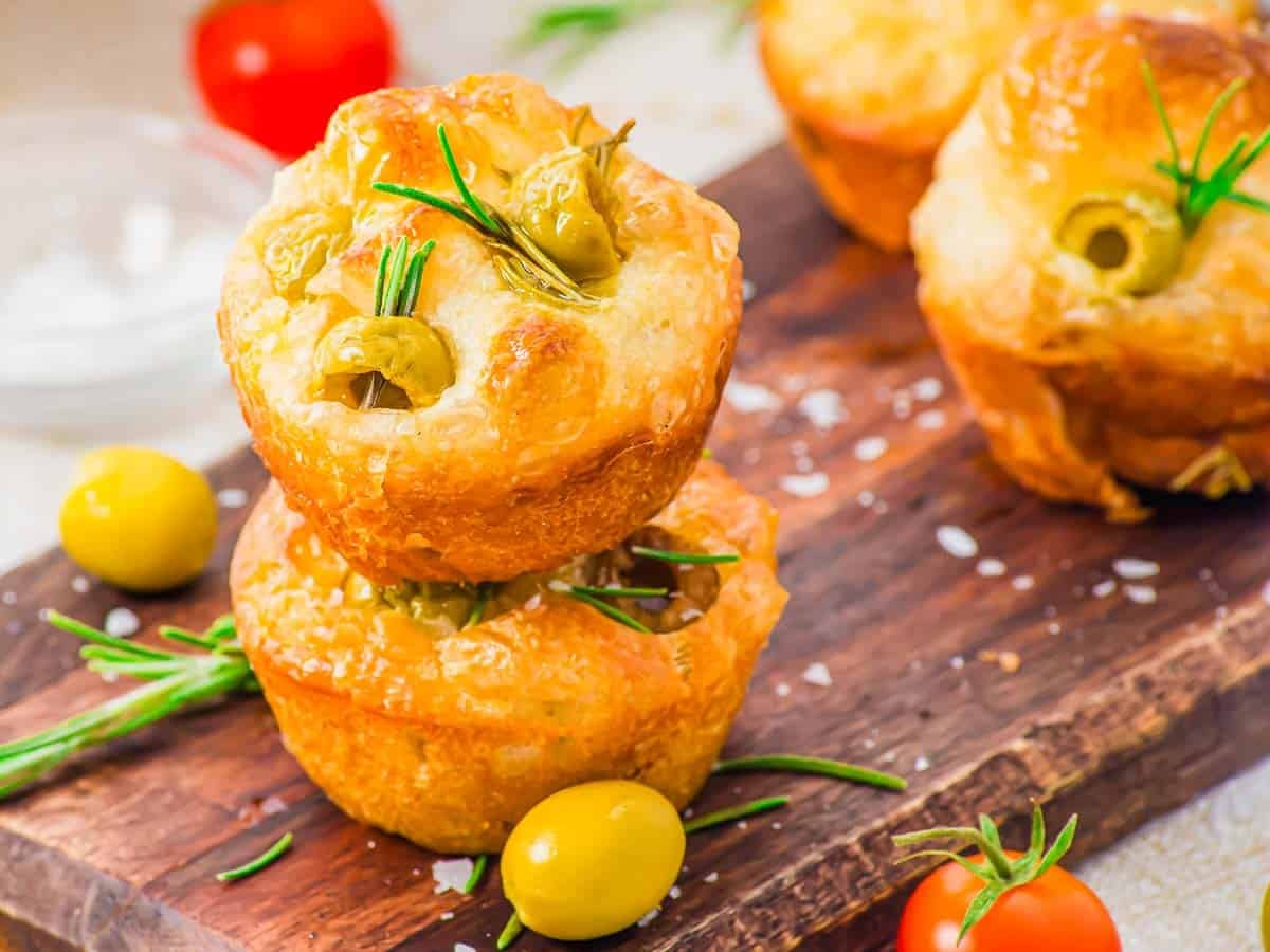 Two savory focaccia muffins topped with green olives and rosemary are stacked on a wooden board, surrounded by whole olives, coarse salt, and cherry tomatoes. More muffins are in the background.