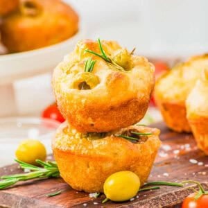 Two focaccia muffins topped with rosemary and green olives are stacked on a wooden board, surrounded by olives, rosemary sprigs, and a sprinkle of sea salt, with more muffins blurred in the background.