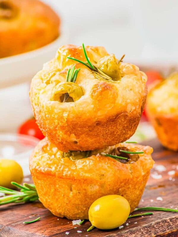 Two focaccia muffins topped with rosemary and green olives are stacked on a wooden board, surrounded by olives, rosemary sprigs, and a sprinkle of sea salt, with more muffins blurred in the background.
