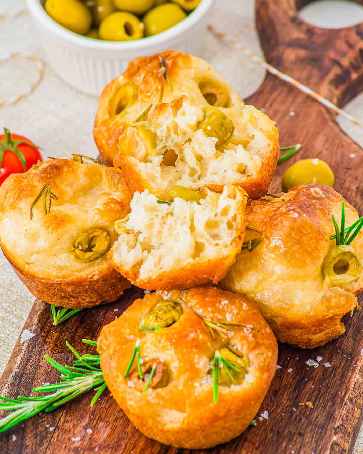 Golden brown olive focaccia muffins topped with green olives and rosemary are stacked on a wooden board, with a small bowl of olives and a sprig of fresh rosemary nearby.