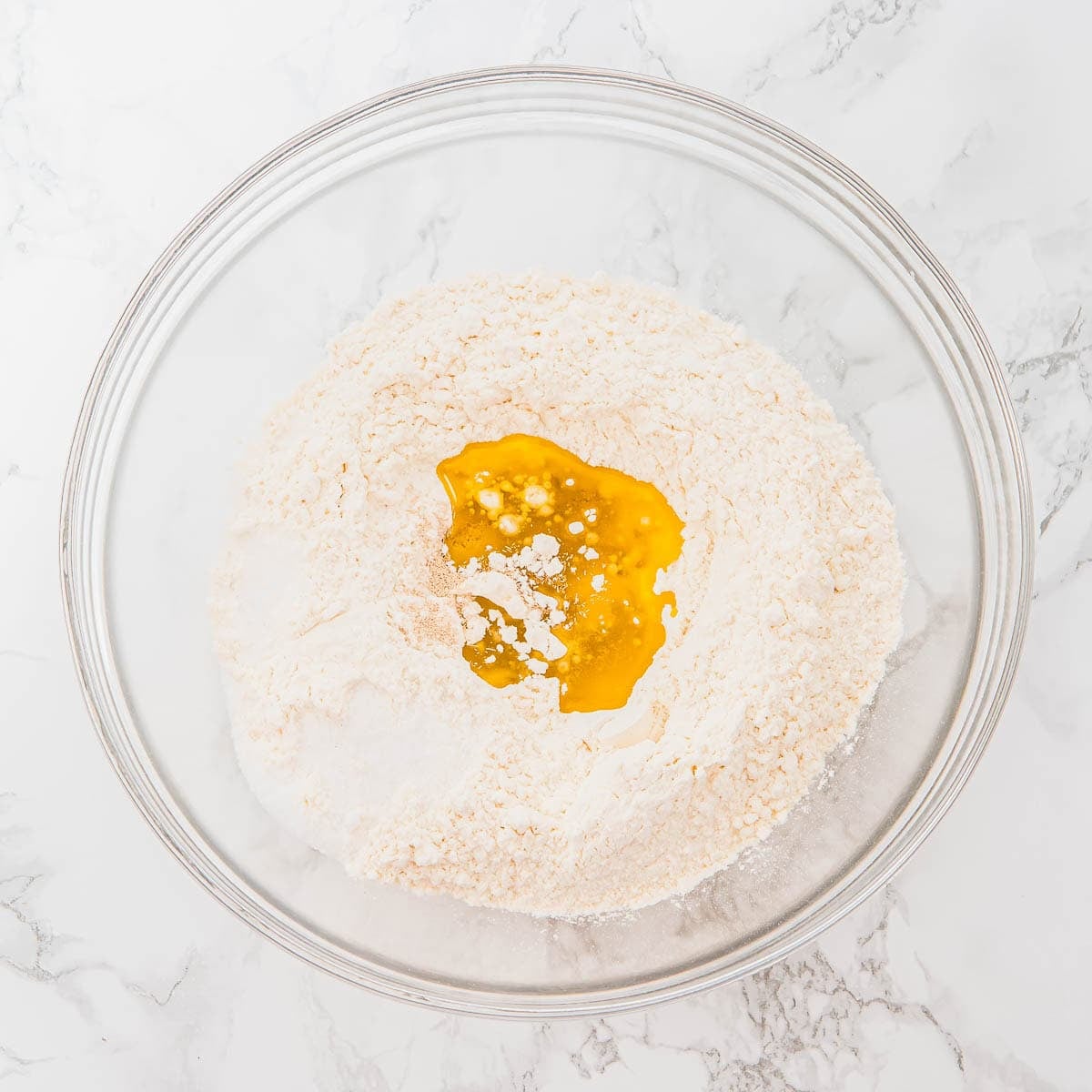 A glass bowl on a marble surface contains flour with olive oil, ready for mixing.