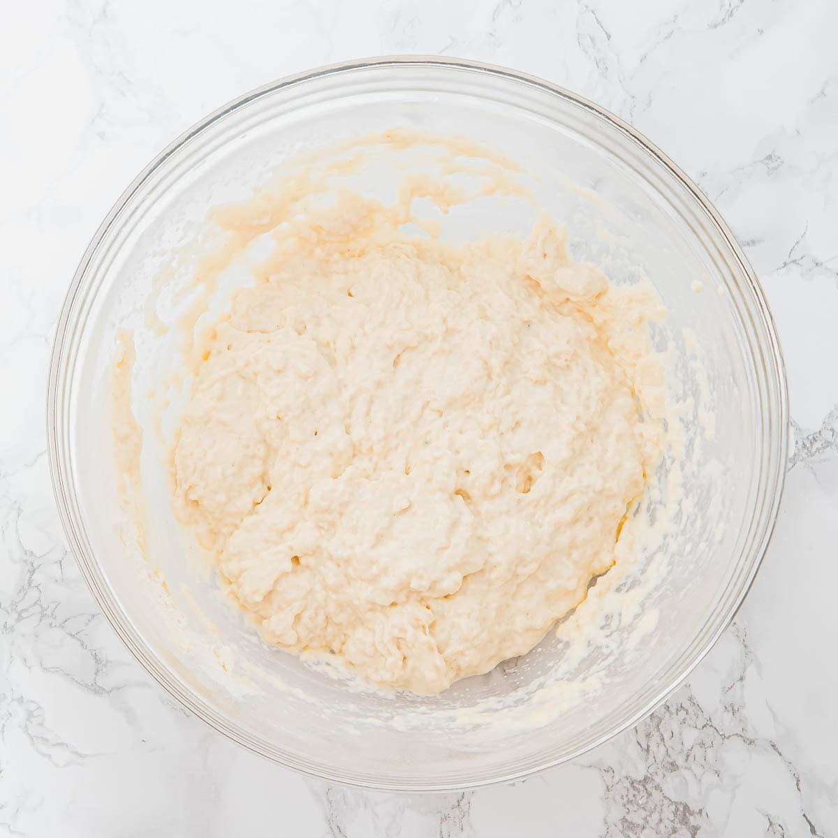 A glass bowl filled with pale, sticky focaccia bread dough sits on a white marble surface.