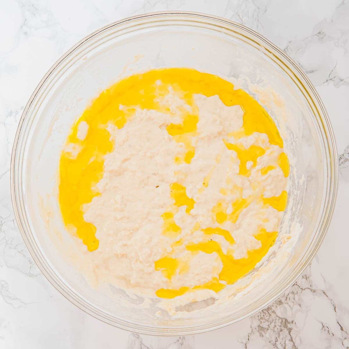 A glass bowl containing focaccia dough with olive oil partially blended, sits on a white marble surface.