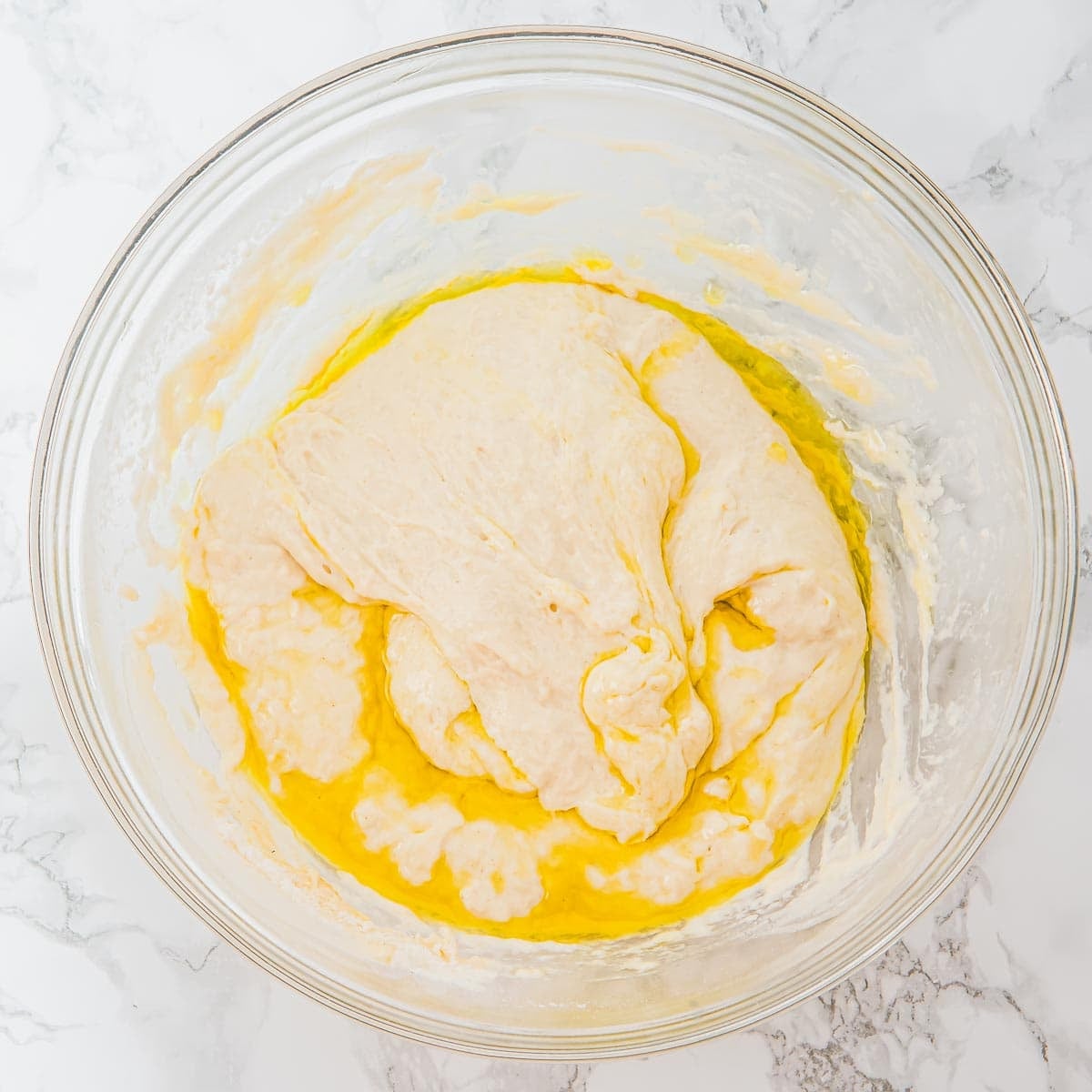 A glass bowl containing focaccia dough partially mixed with olive oil, sitting on a white marble surface.