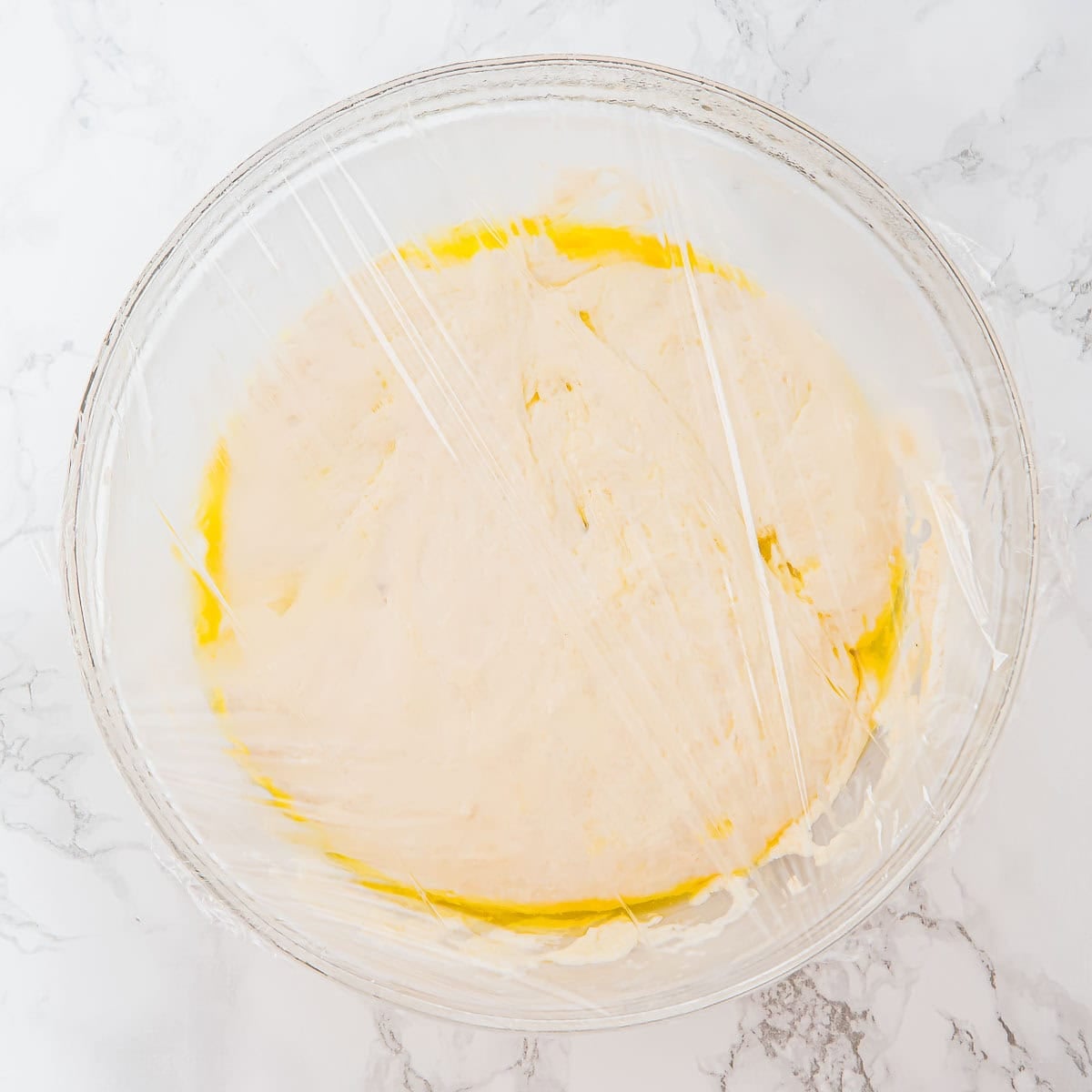 A glass bowl filled with dough covered in plastic wrap, resting on a white marble surface. The dough appears to be rising, with a yellowish tint around the edges.