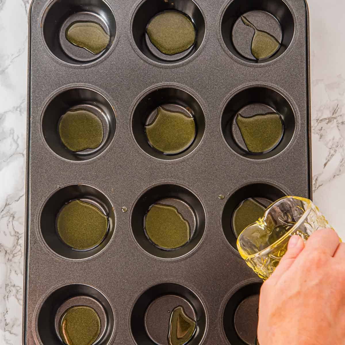 A hand pours oil from a glass into the cups of a 12-cup muffin tin on a white marble surface. Some cups already contain small pools of oil.
