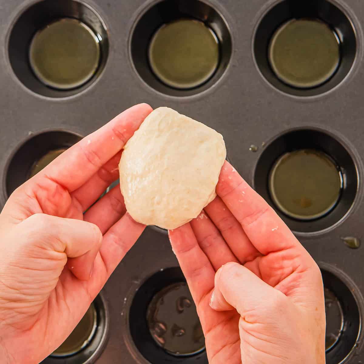 A person holds a piece of focaccia dough over a muffin tin with several cups lightly coated in oil, preparing to place the dough into one of the cups.