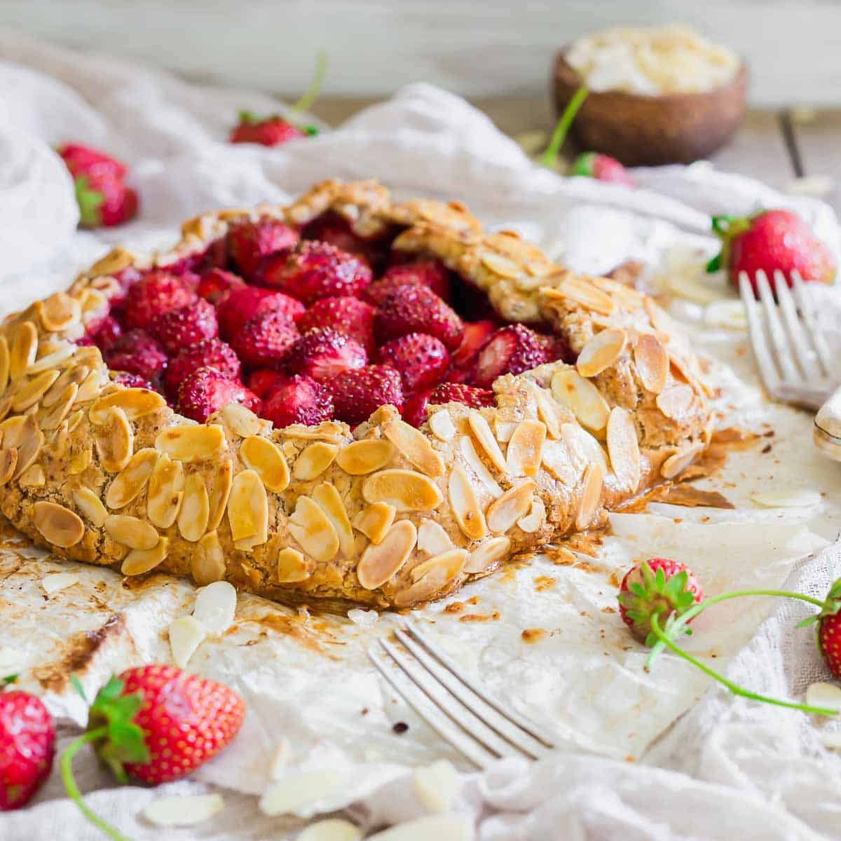 A rustic strawberry galette topped with sliced almonds sits on parchment paper, surrounded by fresh strawberries and two forks. The crust is golden and flaky, with scattered almond slices.