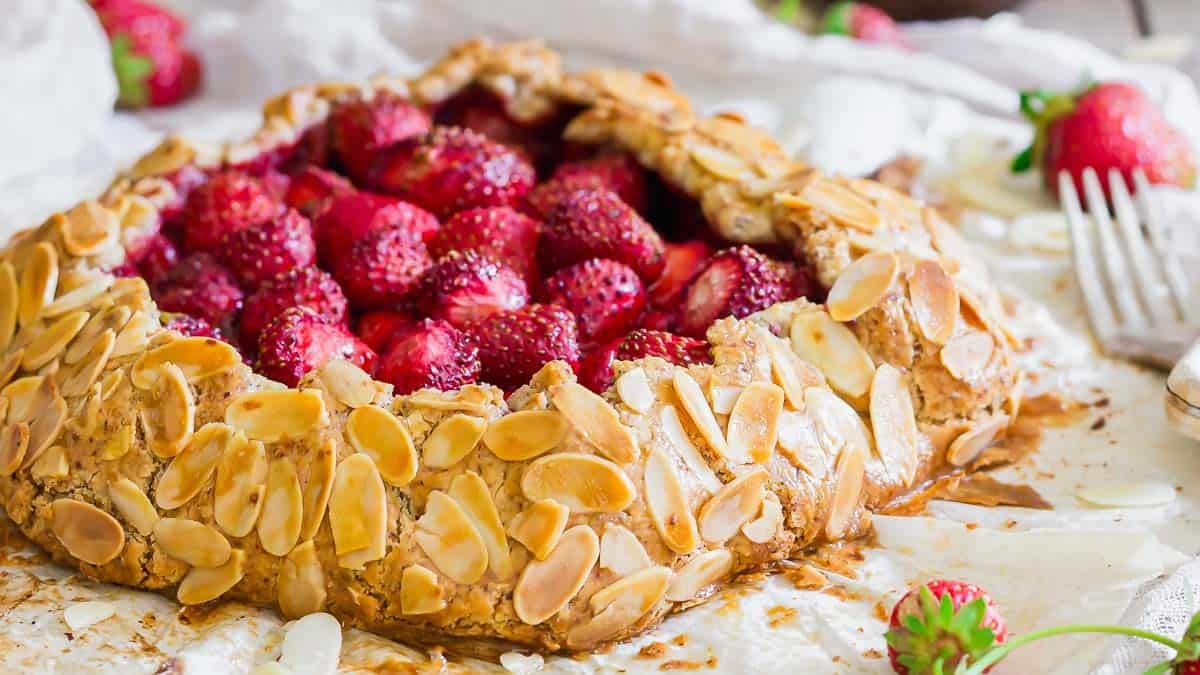 A rustic strawberry galette with a golden almond-crusted edge, filled with fresh strawberries, sits on parchment paper with a fork and loose strawberries nearby.