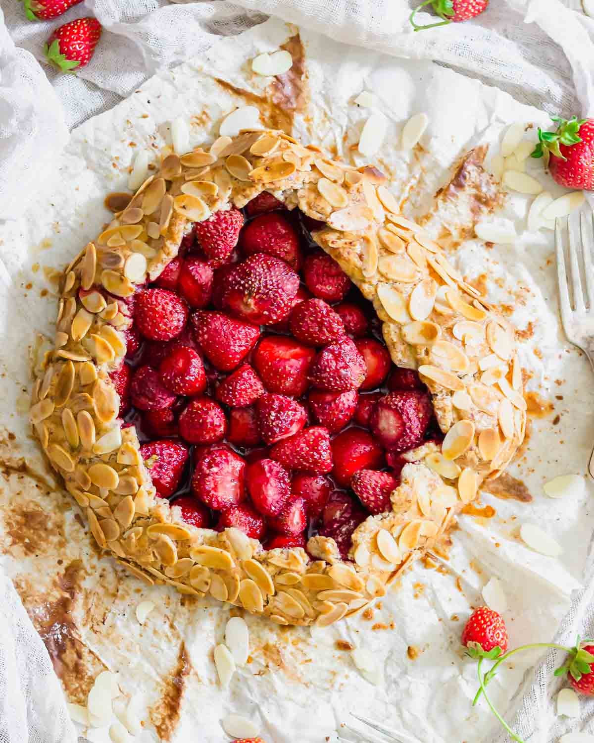 A strawberry galette with a golden almond-crusted crust, filled with fresh strawberries, sits on parchment paper. Sliced almonds and whole strawberries are scattered around. Two silver forks are nearby.