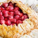 A galette filled with fresh strawberries and topped with sliced almonds sits on parchment paper, with a fork nearby and a few strawberries scattered around.