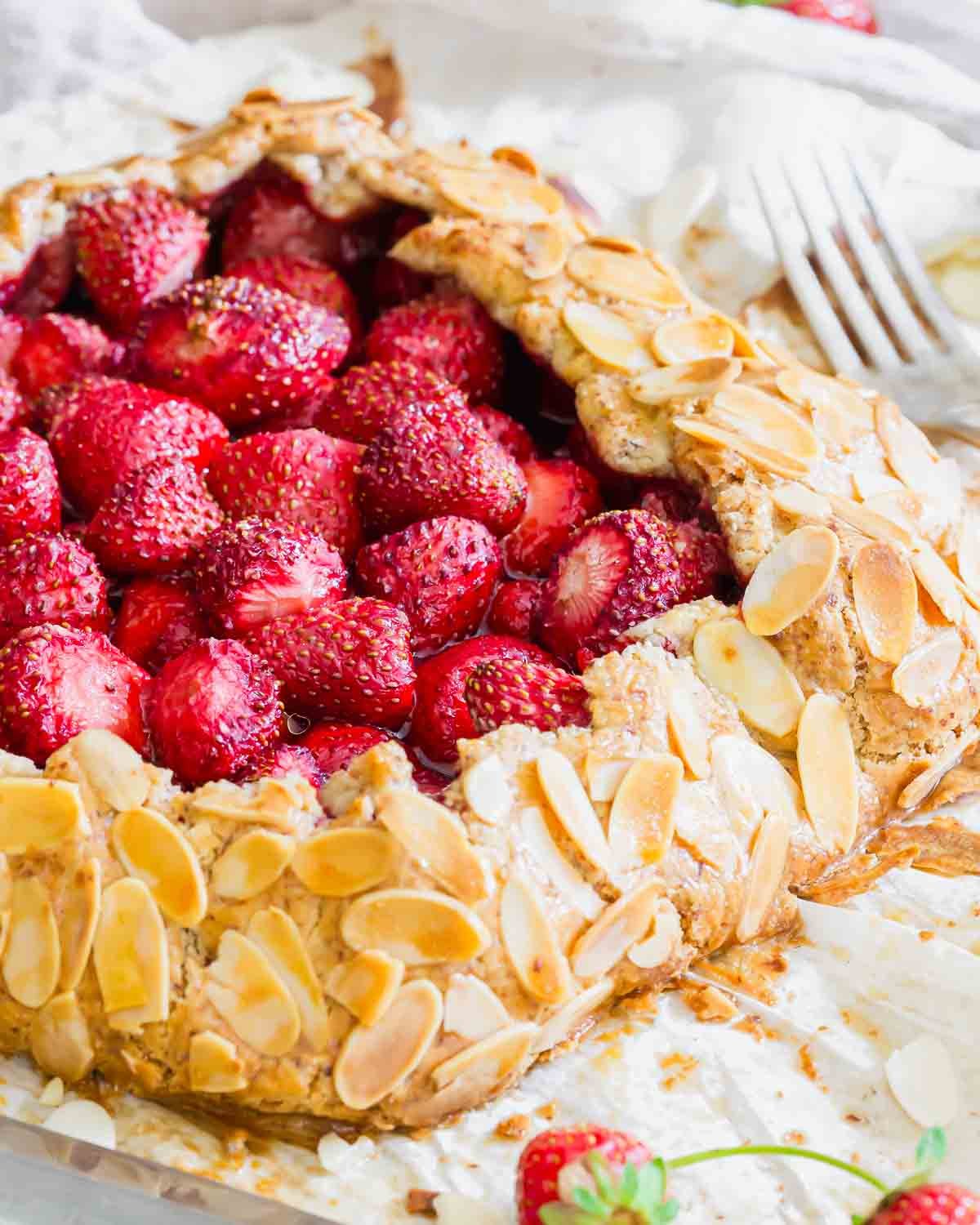 A galette filled with fresh strawberries and topped with sliced almonds sits on parchment paper, with a fork nearby and a few strawberries scattered around.