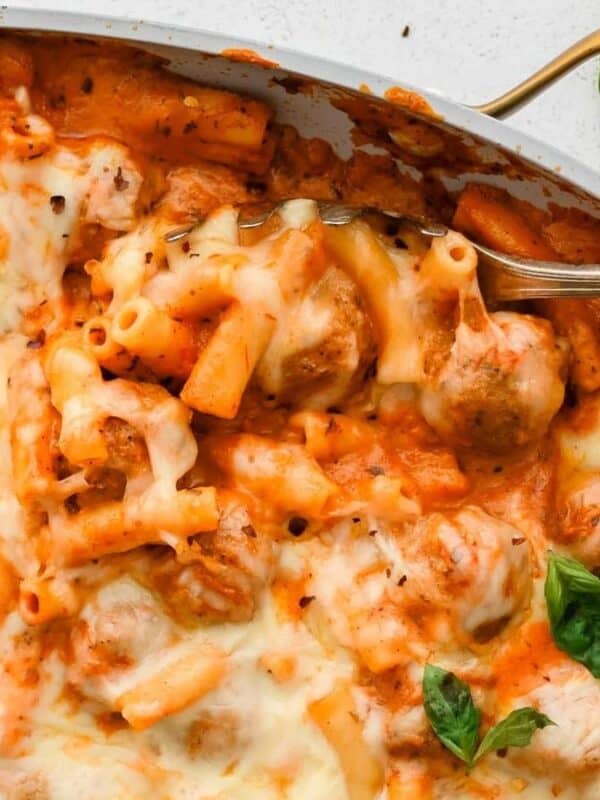 A close-up of baked ziti pasta with melted cheese, tomato sauce, meatballs, and fresh basil leaves in a casserole dish. A spoon is serving a portion from the dish.