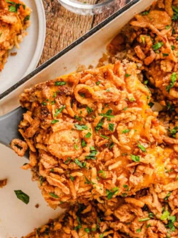 A baking dish filled with a golden-brown casserole topped with crispy fried onions and chopped herbs, with a serving being lifted out on a spatula and a portion plated nearby.