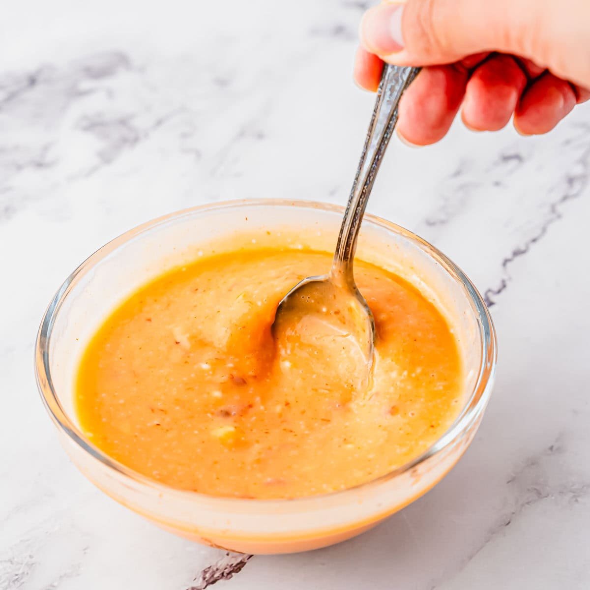 A hand holding a spoon is stirring a bowl of bang bang sauce on a white marble surface.