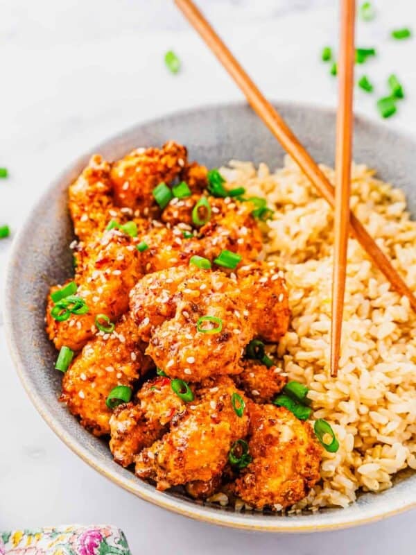 A bowl filled with golden, crispy air fried bang bang shrimp topped with chopped green onions, served next to a portion of brown rice. Wooden chopsticks rest on the bowl’s rim.