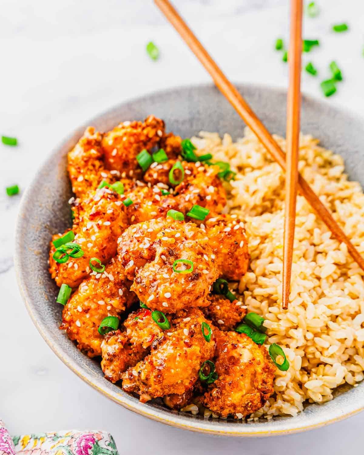 A bowl filled with golden, crispy air fried bang bang shrimp topped with chopped green onions, served next to a portion of brown rice. Wooden chopsticks rest on the bowl&rsquo;s rim.