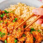 A hand using chopsticks to pick up crispy, saucy bang bang shrimp made in the air fryer garnished with sesame seeds and chopped green onions, served with a side of brown rice in a bowl.
