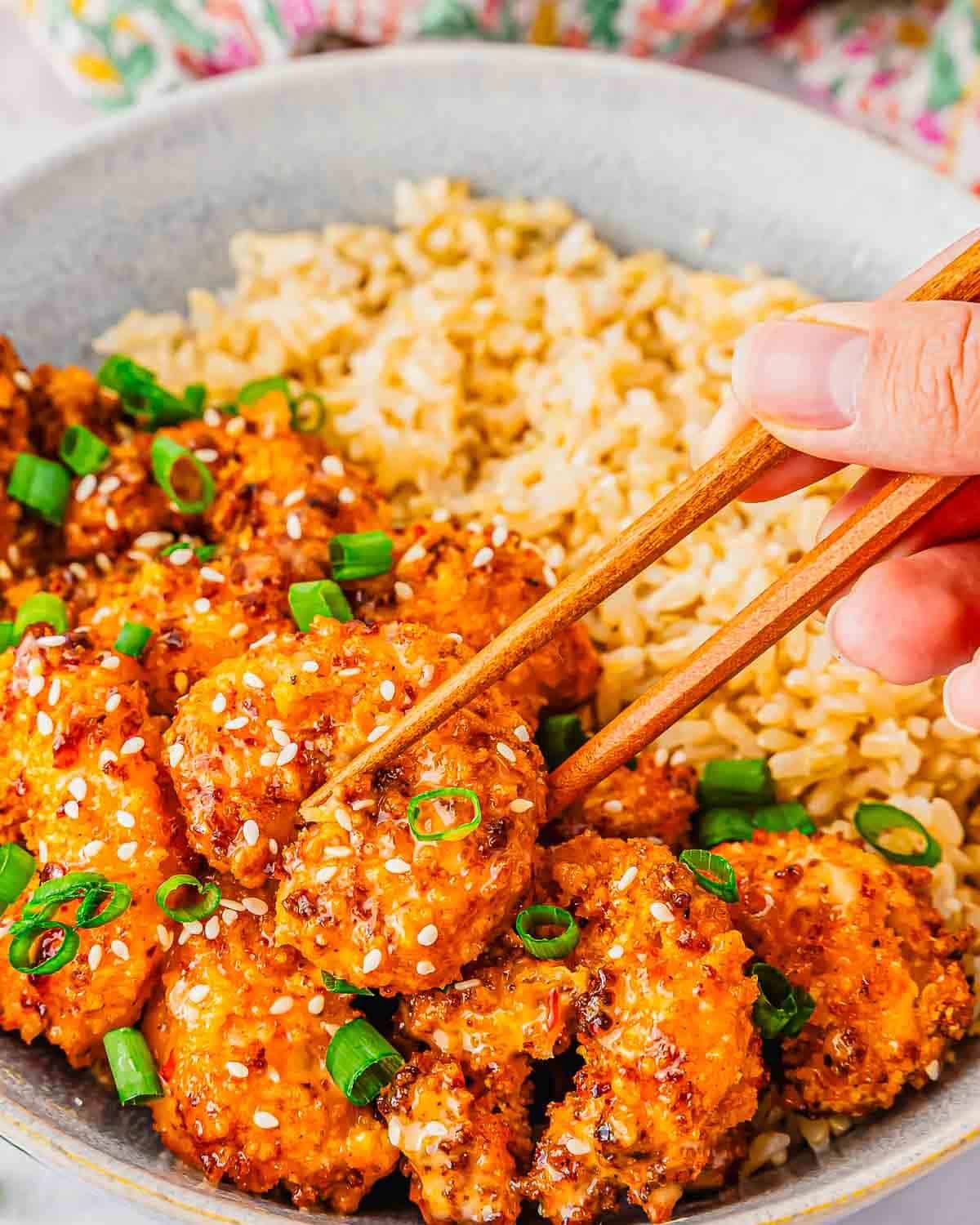 A hand using chopsticks to pick up crispy, saucy bang bang shrimp made in the air fryer garnished with sesame seeds and chopped green onions, served with a side of brown rice in a bowl.