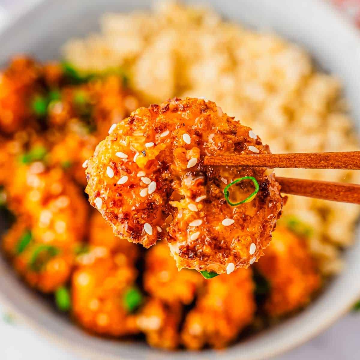 A close-up of chopsticks holding a piece of crispy air fryer bang bang shrimp topped with sesame seeds and green onion, with a bowl of more cauliflower and rice in the blurred background.