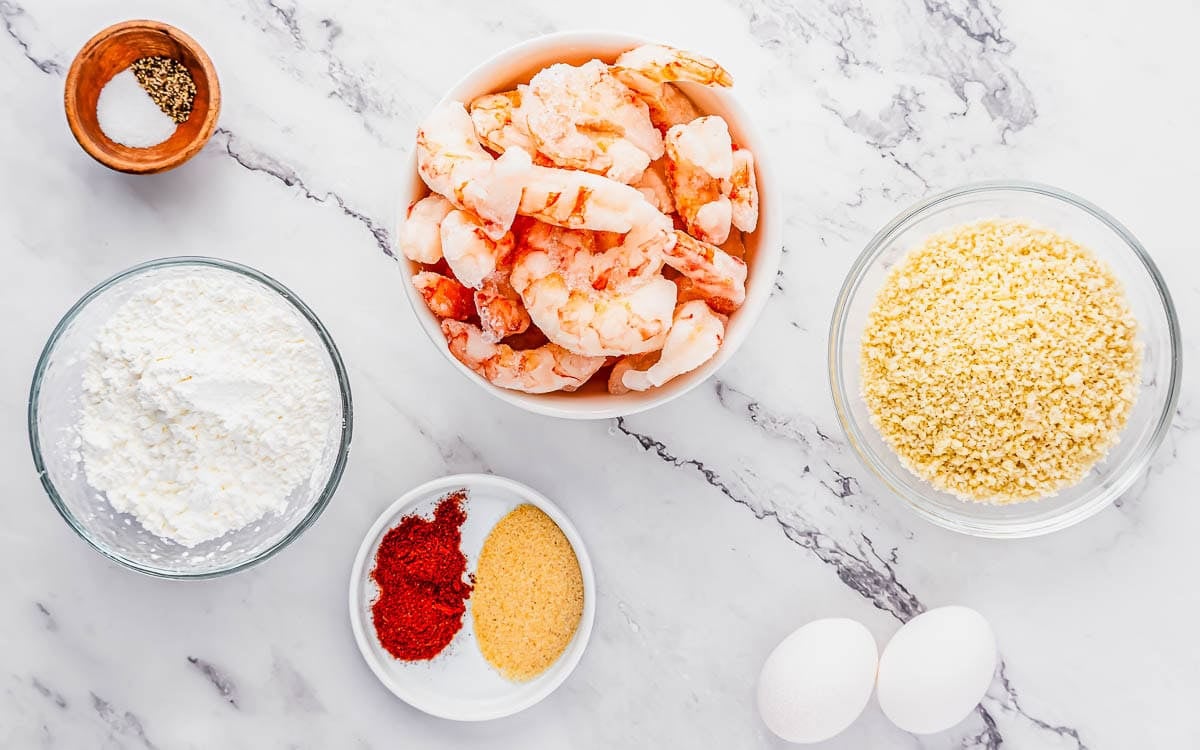 Five bowls and plates containing peeled shrimp, cornstarch, breadcrumbs, spices, salt, and pepper, along with two eggs, are arranged on a white marble surface, ready for meal preparation.