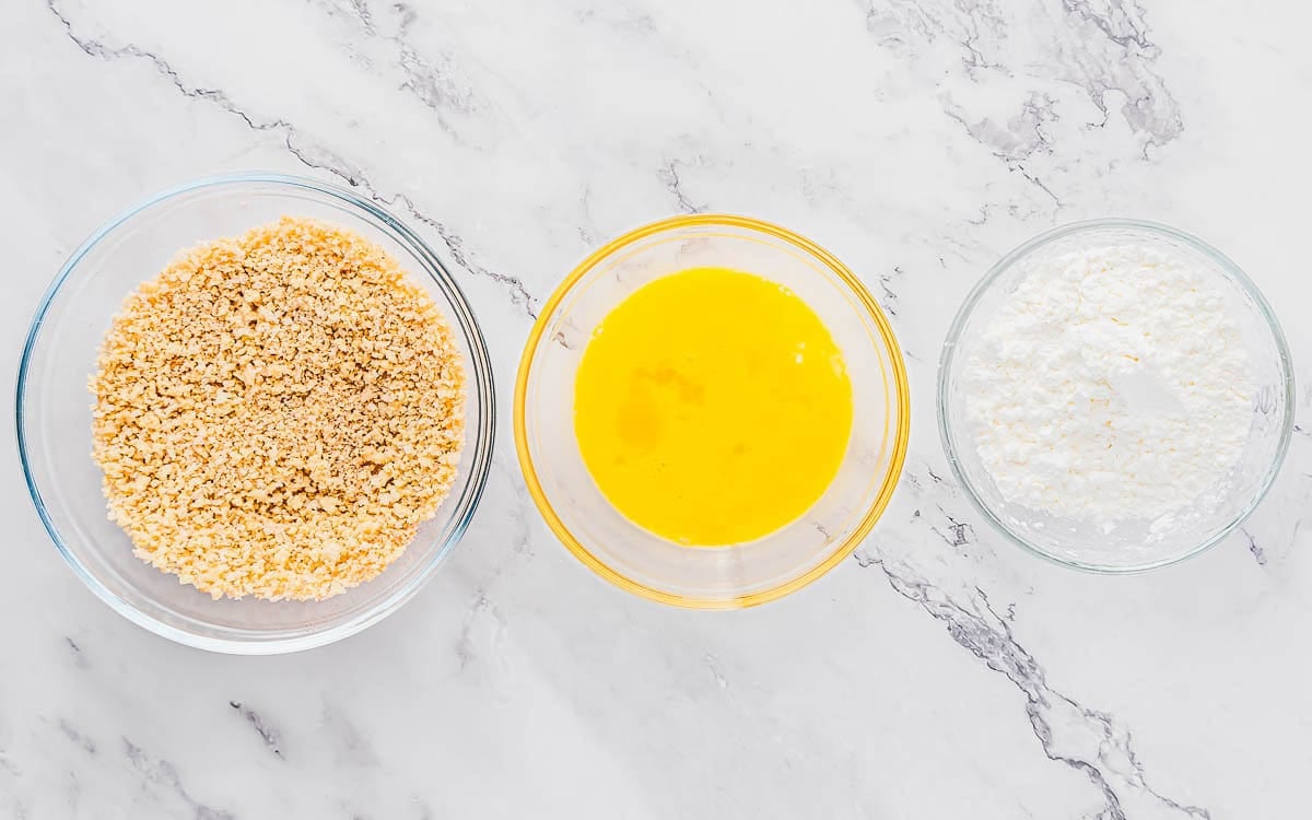 Three glass bowls on a marble countertop: the left bowl contains breadcrumbs, the middle bowl has beaten eggs, and the right bowl holds cornstarch for dredging shrimp.