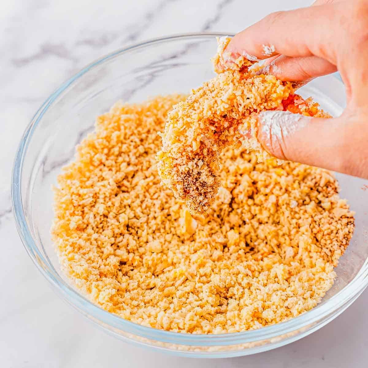 A hand dips shrimp into a bowl of breadcrumbs for coating. The bowl is clear glass and the background is a white marble surface.