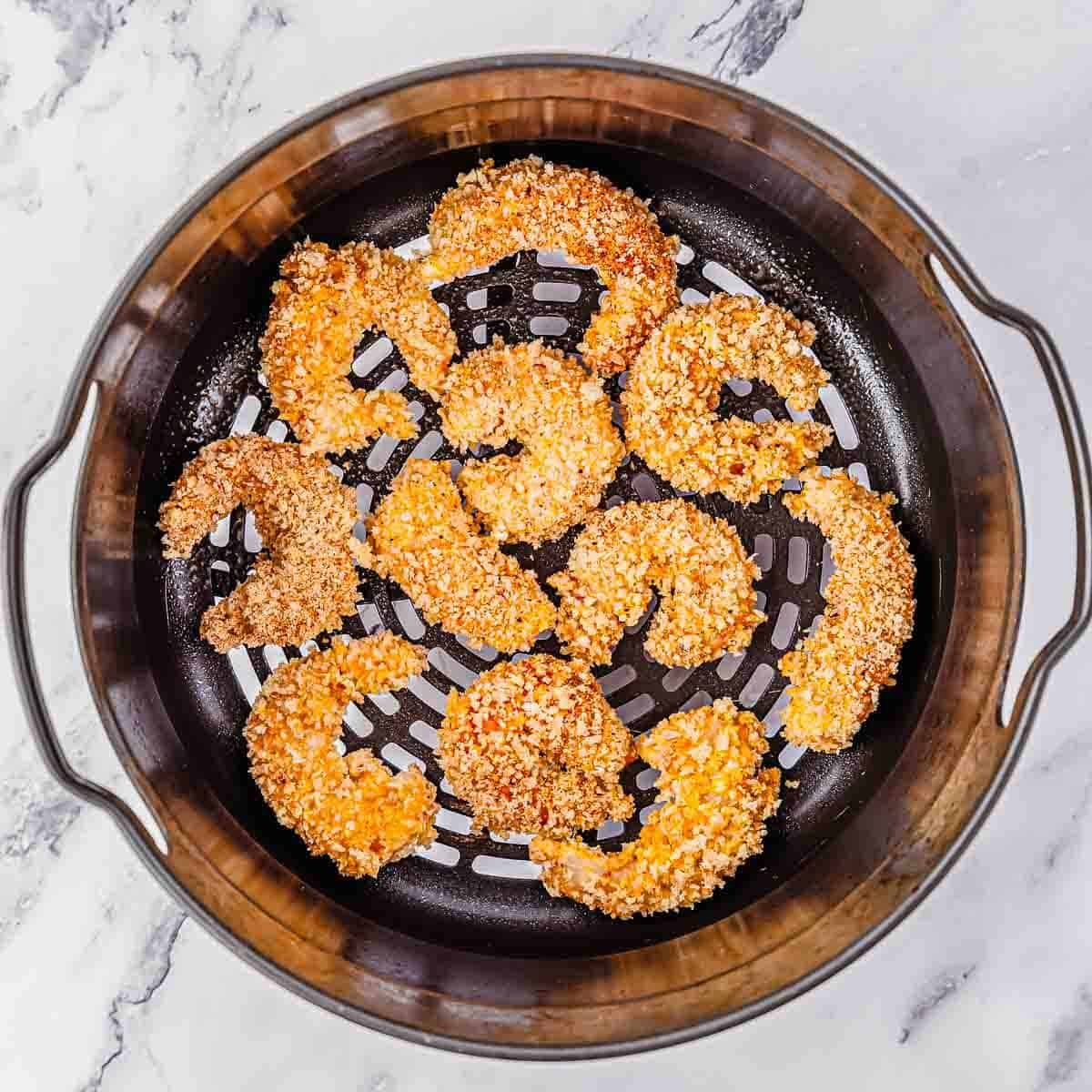 Breaded shrimp arranged in a single layer inside an air fryer basket, ready to be cooked. The background is a white marble surface.