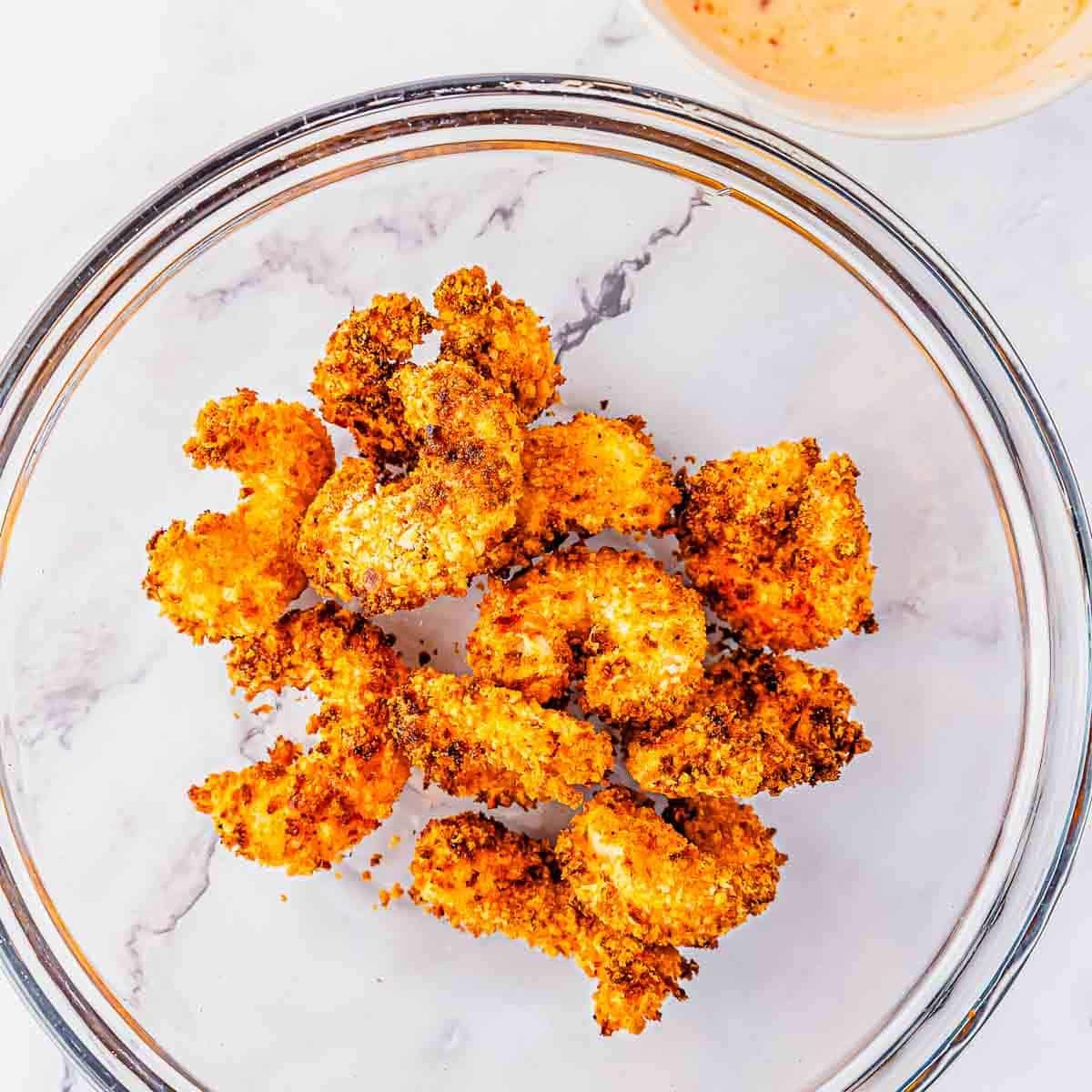 Crispy, golden-brown air fried shrimp arranged in a clear glass bowl on a white marble surface, with a dipping sauce visible in the corner.
