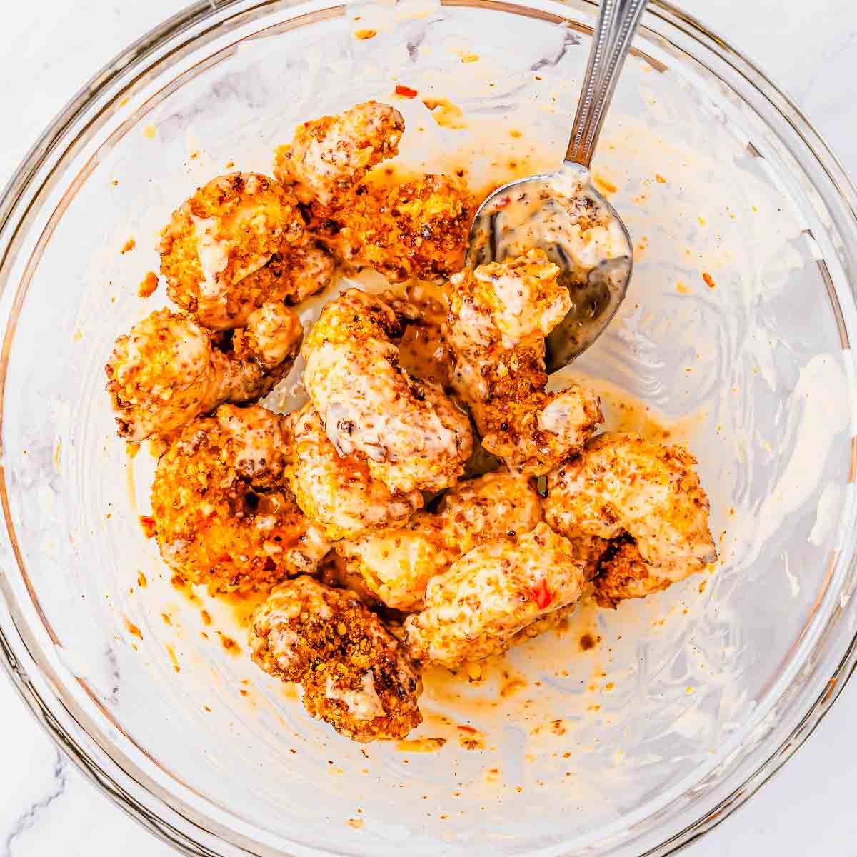 A glass bowl with shrimp coated in a creamy, bang bang sauce. A metal spoon rests inside the bowl, partially covered in the marinade. The bowl sits on a white surface.