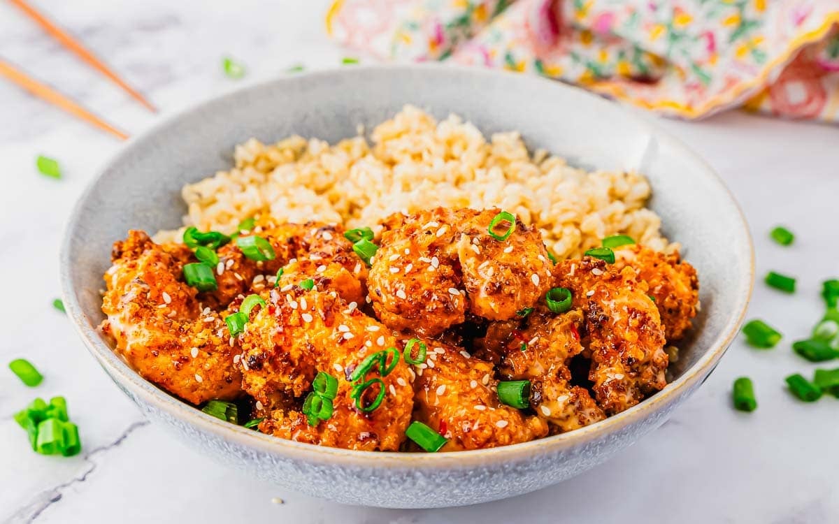A bowl of crispy bang bang shrimp bites topped with sesame seeds and chopped green onions, served alongside a portion of brown rice.