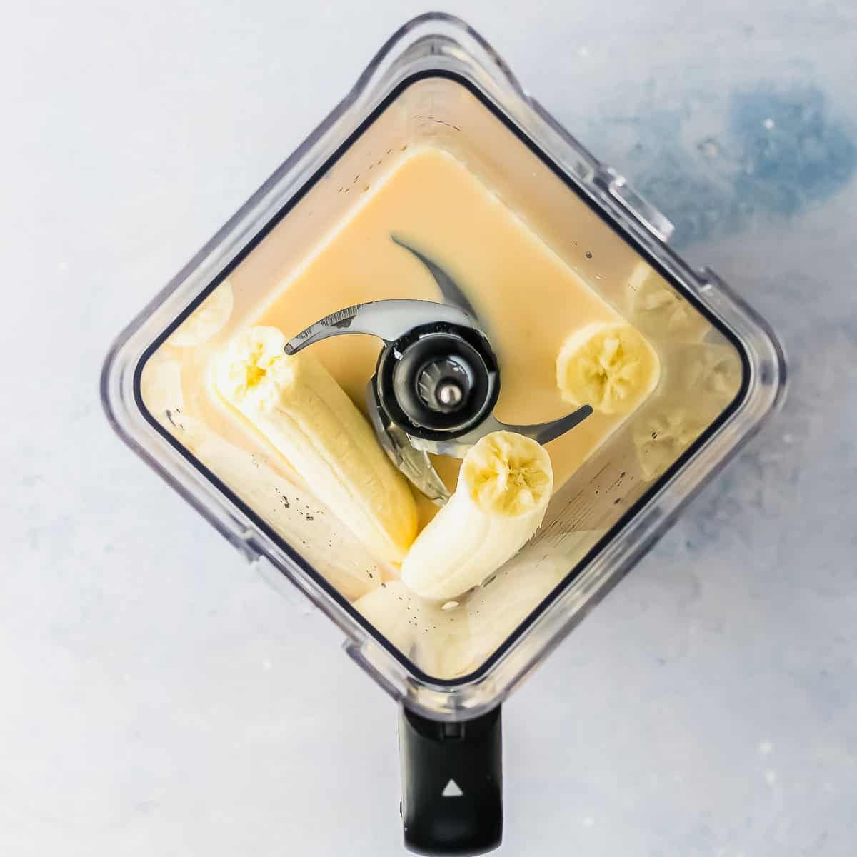 Top view of a blender containing peeled bananas and a liquid, ready to be blended, on a light blue surface.