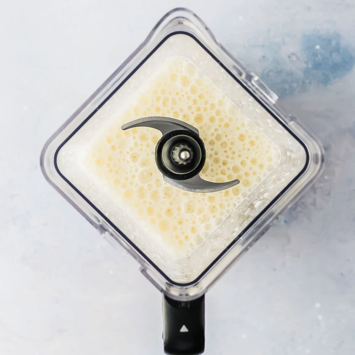 A top-down view of a blender filled with a frothy, creamy banana milk liquid, blending on a light-colored surface.